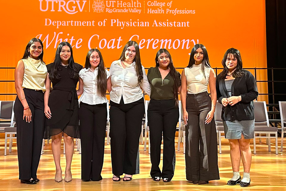 Seven people stand side by side on a stage during a White Coat Ceremony, with an orange backdrop and rows of chairs behind them.