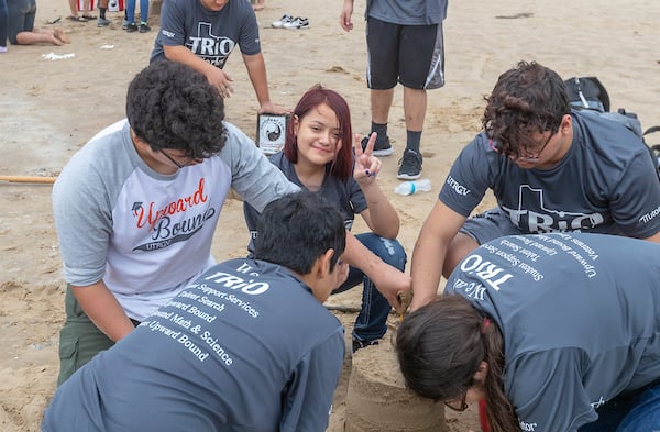 High school student gives V's up while other Upward Bound students work on sandcastle