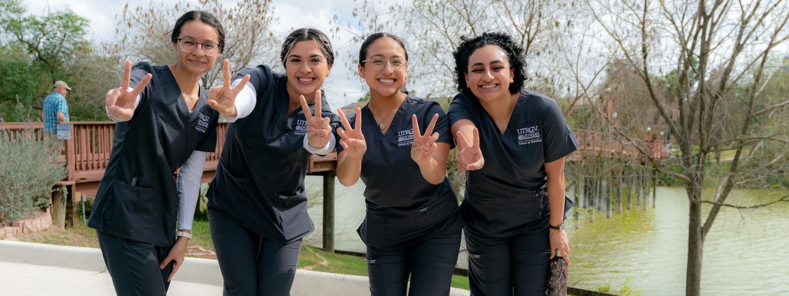 Students in gray scrubs signaling v's up outdoors.