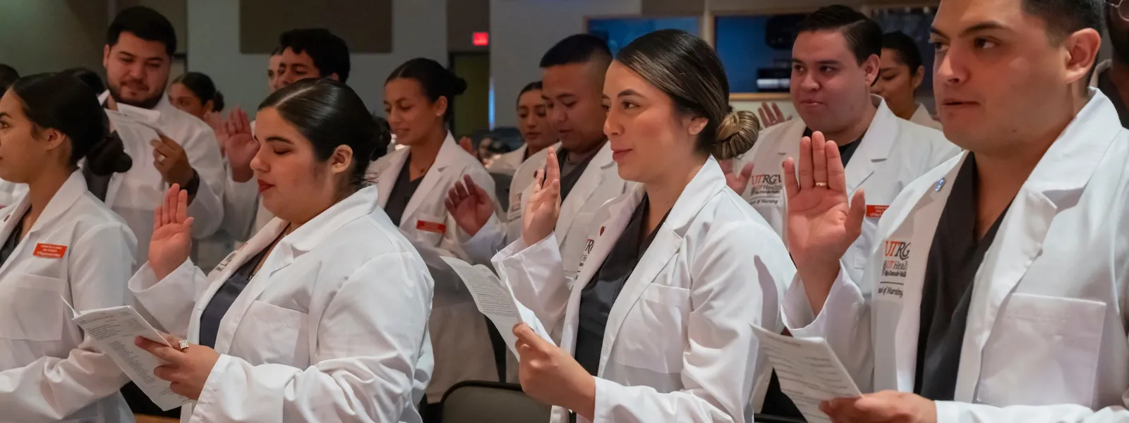 Group of UTRGV nursing students in white coats standing with raised hands while reciting the professional pledge.