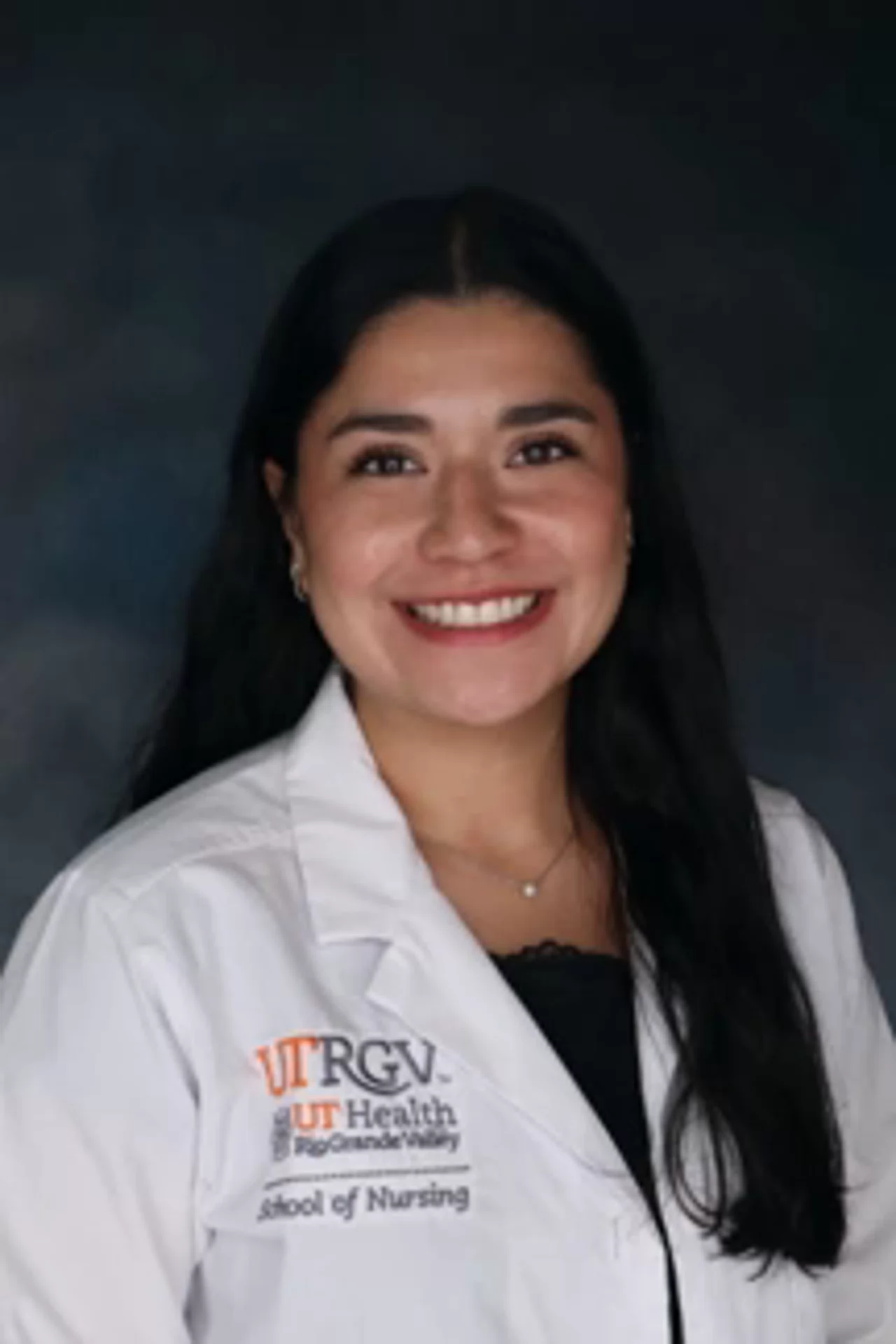 Portrait of Nicole Cuellar wearing a white UTRGV School of Nursing lab coat, smiling at the camera against a dark studio background.