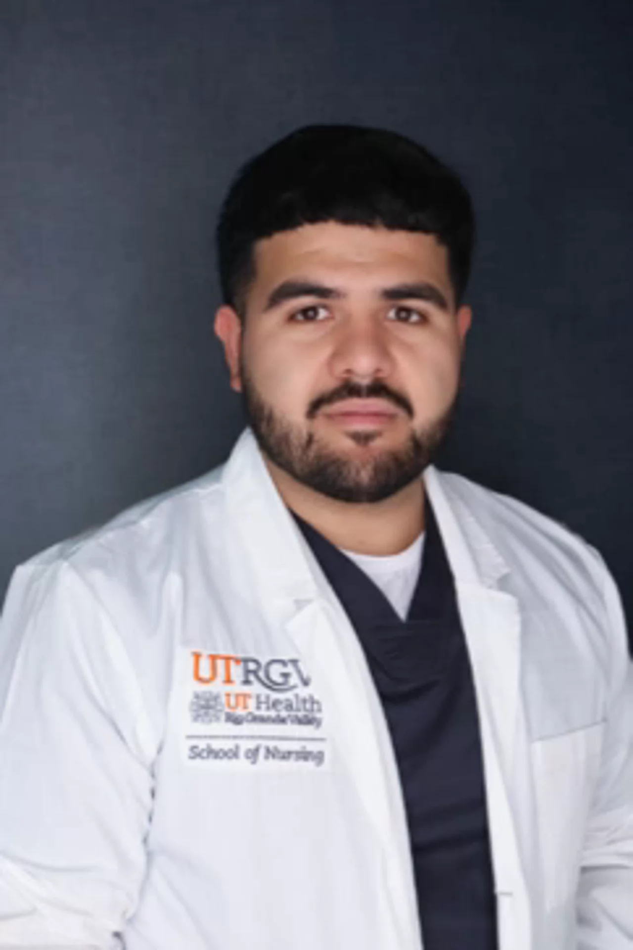 Portrait of Raul Sifuentes Jr wearing a white UTRGV School of Nursing lab coat, facing the camera against a dark background.