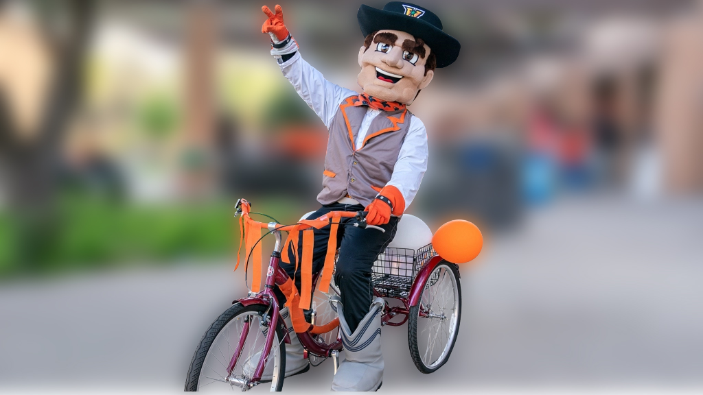 Vaquero Mascot rides a tricycle.