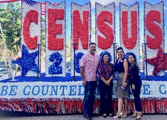 Four individuals stand in front of a red, white, and blue float that reads “Census 2020” and “Be Counted.”