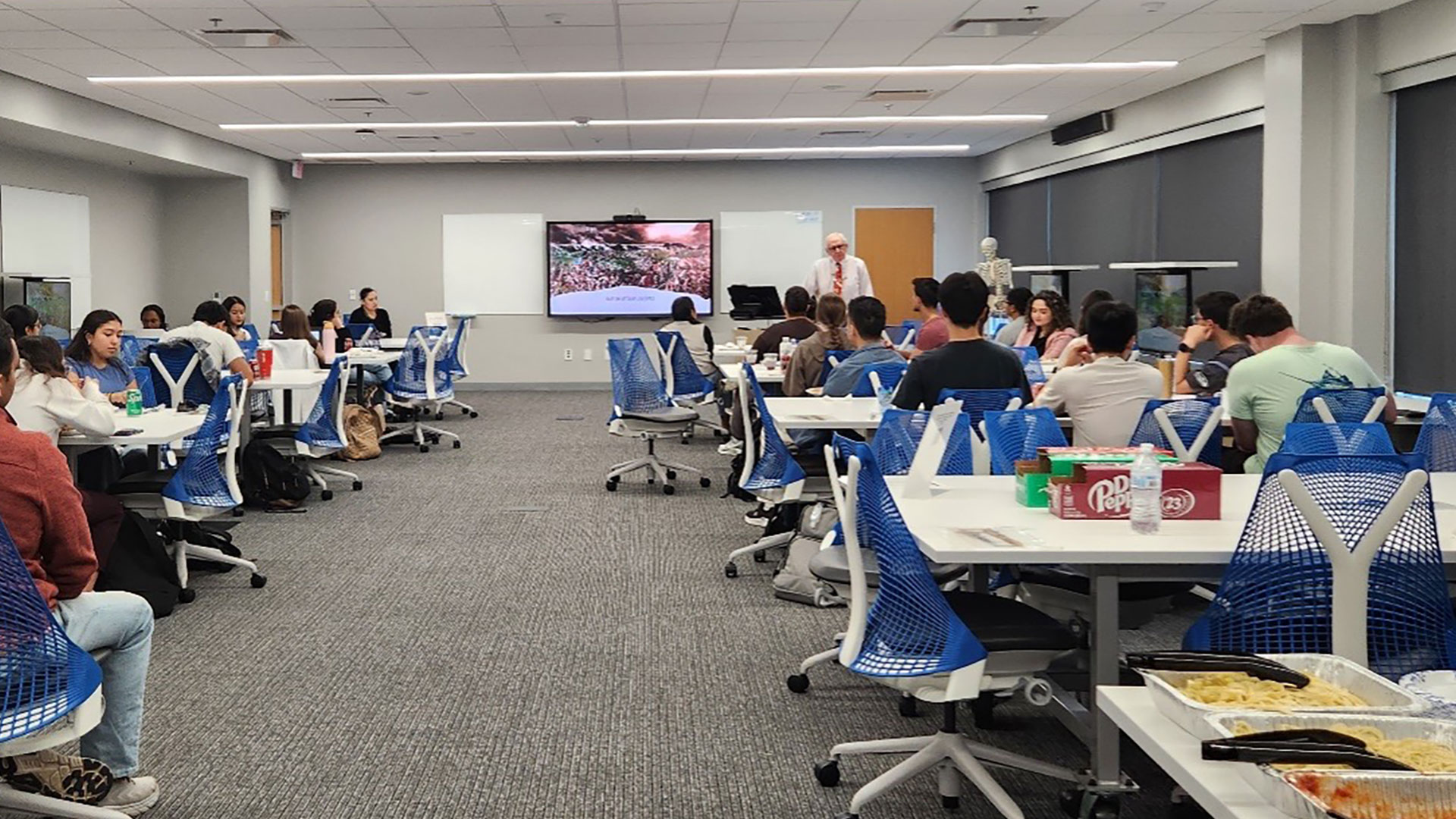 A large group of students sits at tables in a modern classroom, listening to a lecturer who is presenting on a large screen.