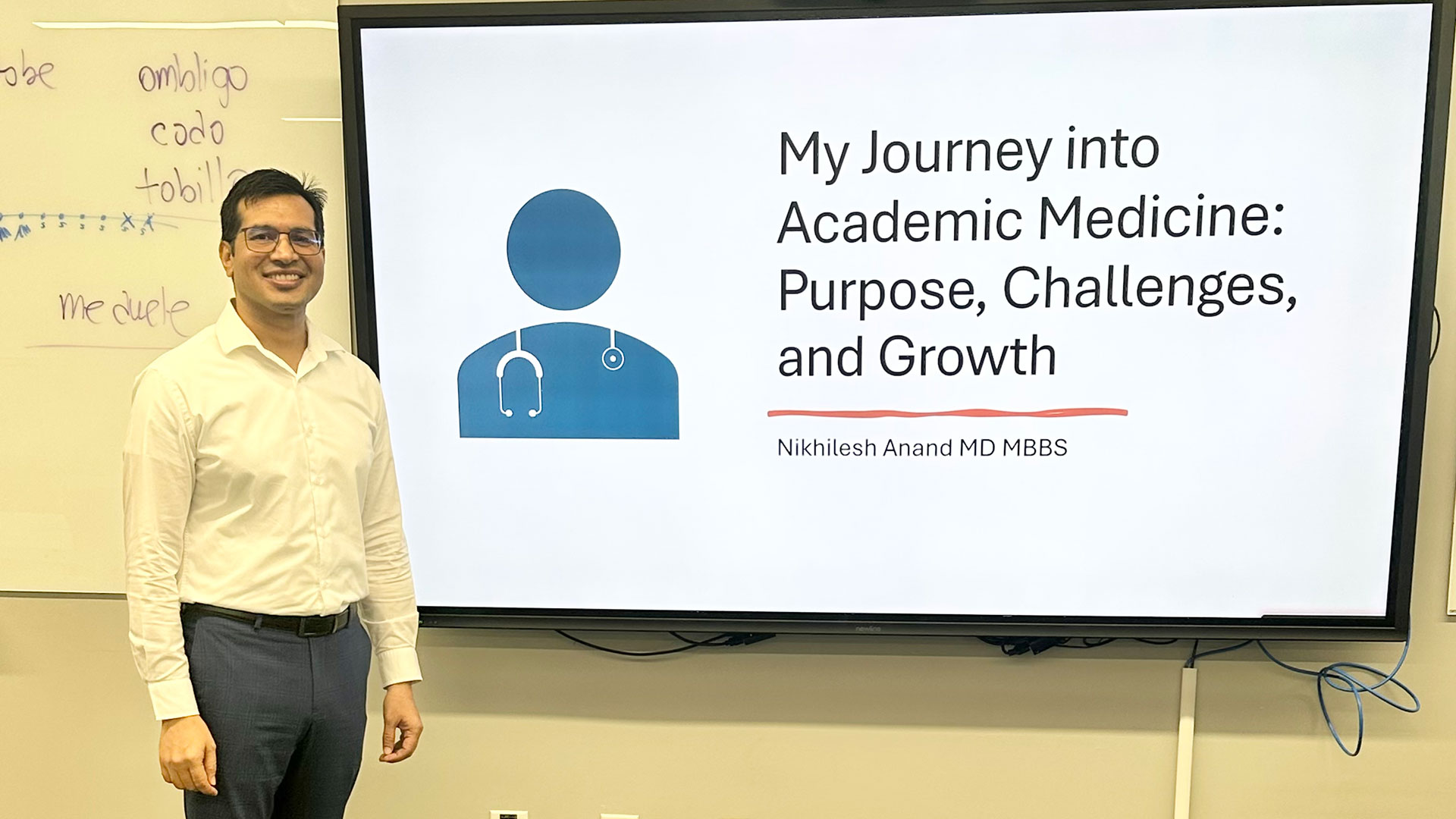 A smiling man stands in front of a presentation screen titled "My Journey into Academic Medicine: Purpose, Challenges, and Growth" by Nikhilesh Anand MD MBBS.