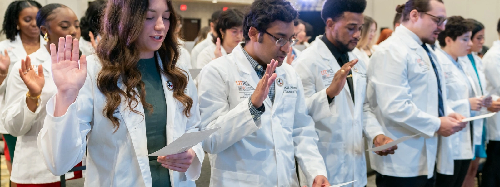 A row of students in white coats recite the professional oath, holding papers and raising their right hands.