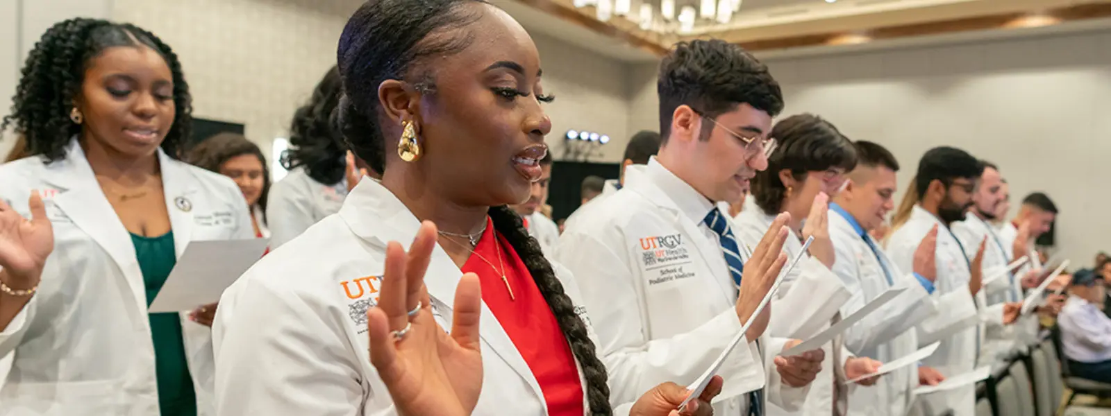 SOPM students in white coats raising their right hands to take an oath during a ceremony. 