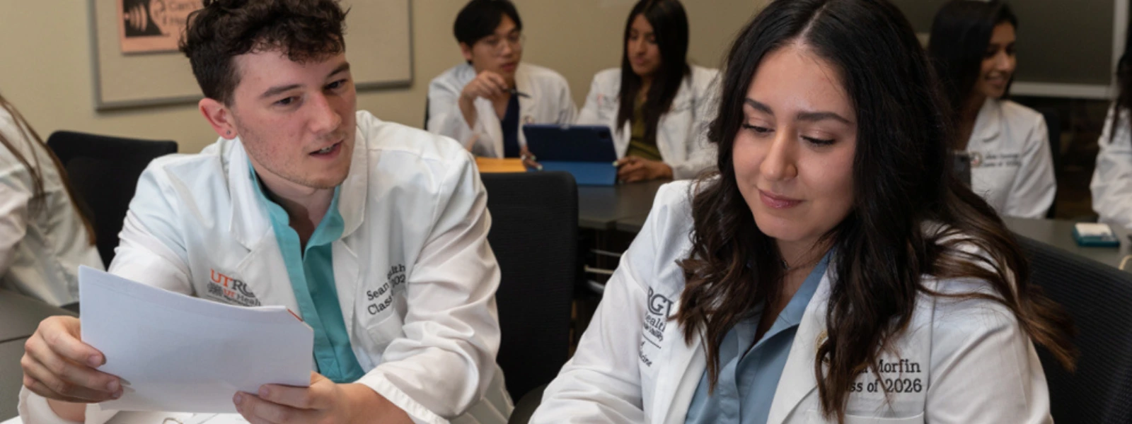 Two SOPM students seated in a classroom reviewing a laptop screen and paperwork together.