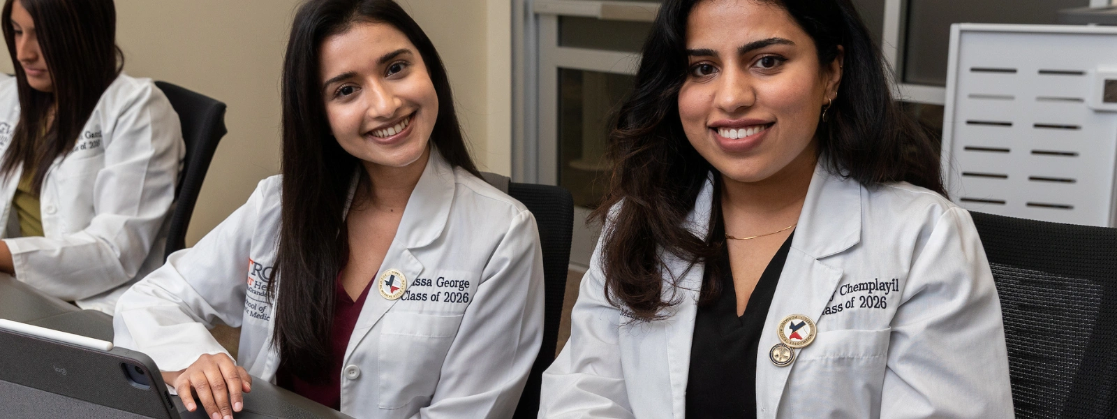 Two SOPM students smiling at camera while sitting in classroom desk.