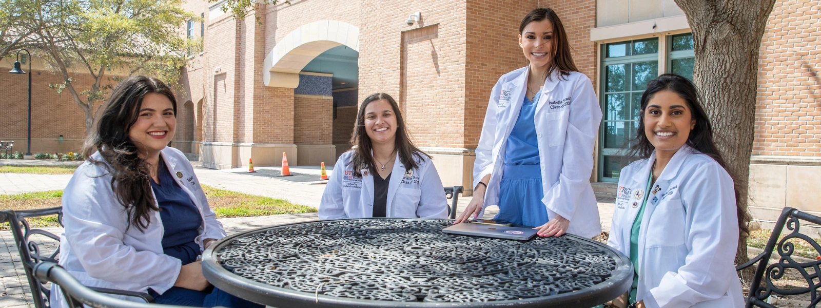 SOPM students in white coats outdoors, one standing and others seated, smiling.