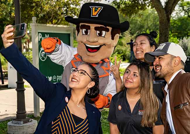 A group of students pose for a selfie with a school mascot outdoors on campus.
