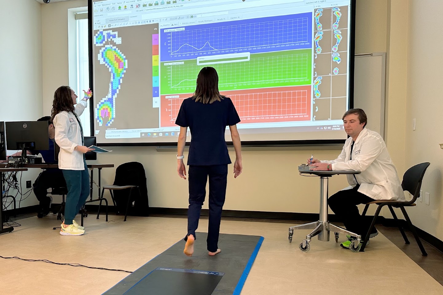 SOPM student facing class screen while one SOPM student watches patient and one SOPM student examines screen.