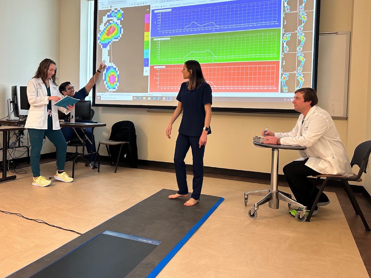 Patient standing in center of room while SOPM students in white coats watch.