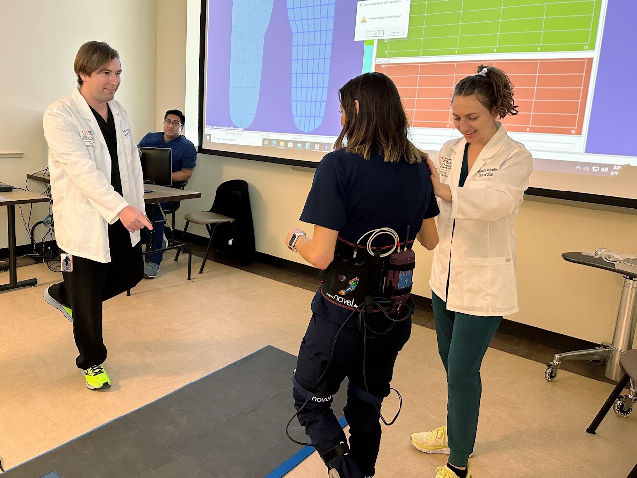 SOPM students in white coats helping patient walk. 
