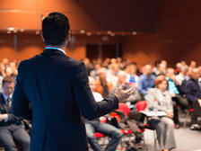 Person in a suit, with back to the camera, presenting onstage during an event or conference.