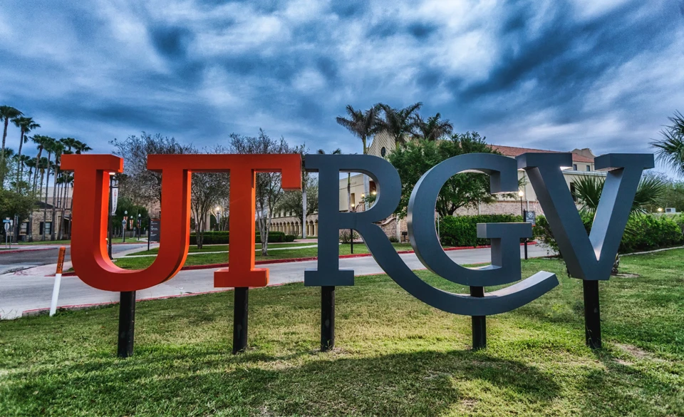 Large UTRGV campus sign displayed outdoors with palm trees and university buildings in the background under a cloudy sky.