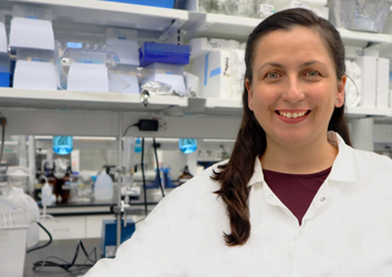 Dr. pereira a woman with dark brown long hair, and blue eyes, poses smiling wearing a white lab coat  with her lab as her background