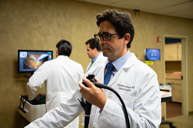 A doctor holds a medical instrument while looking at a monitor; two other doctors are in the background.