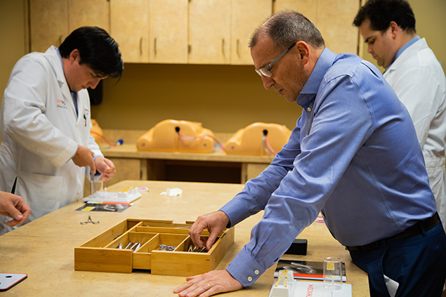 Three people are shown in a medical training classroom, working with surgical instruments and anatomical models.