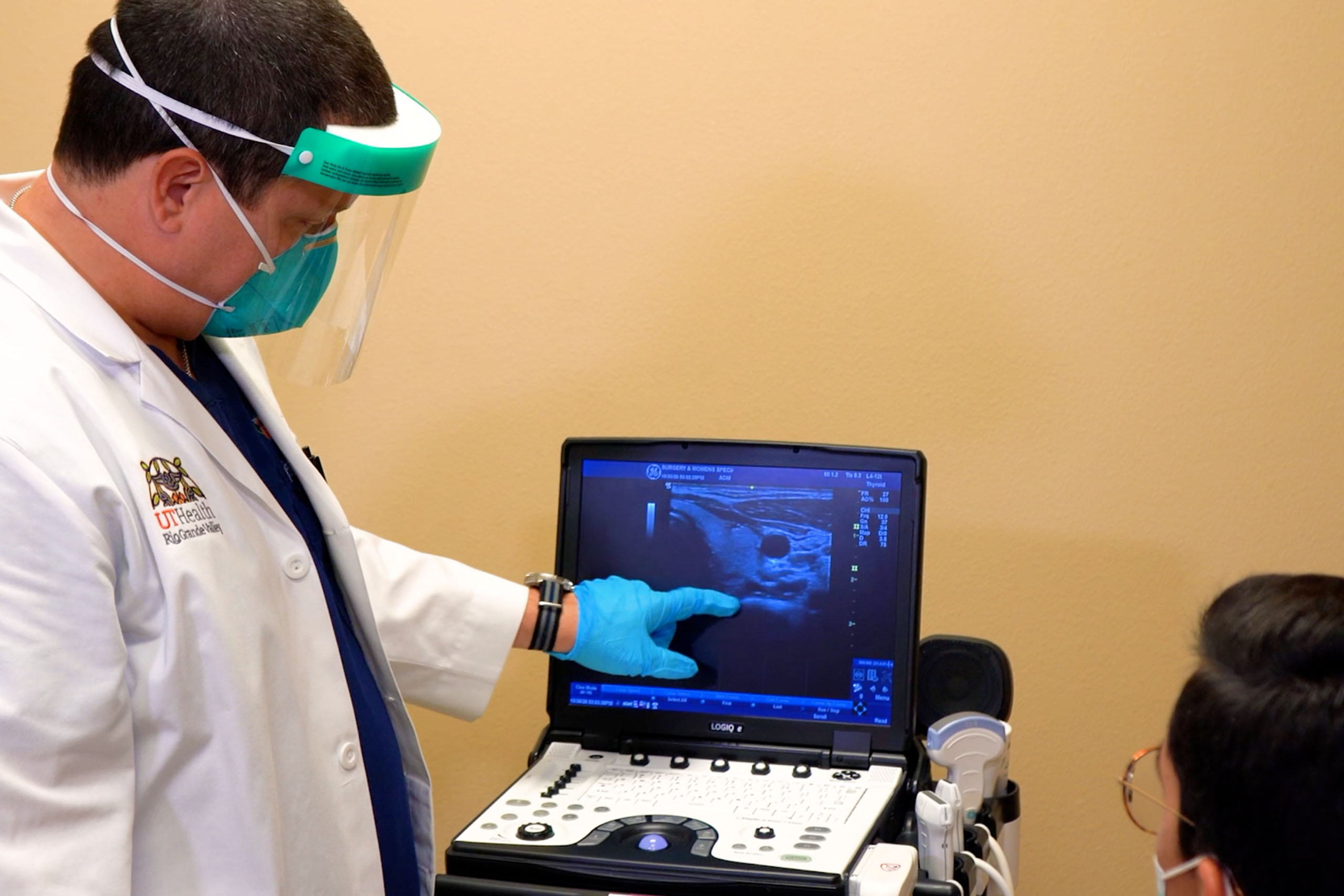 A healthcare worker wearing full PPE including a face shield, mask, gloves, and gown points at an ultrasound image on a portable machine being viewed by a patient in a mask.