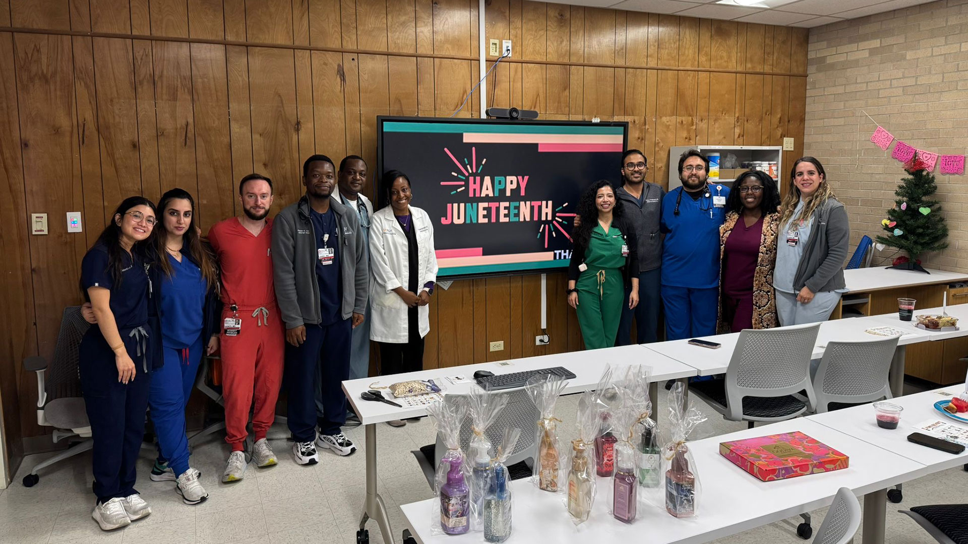 A group of healthcare workers pose for a photo in front of a screen displaying a "Happy Juneteenth" message.