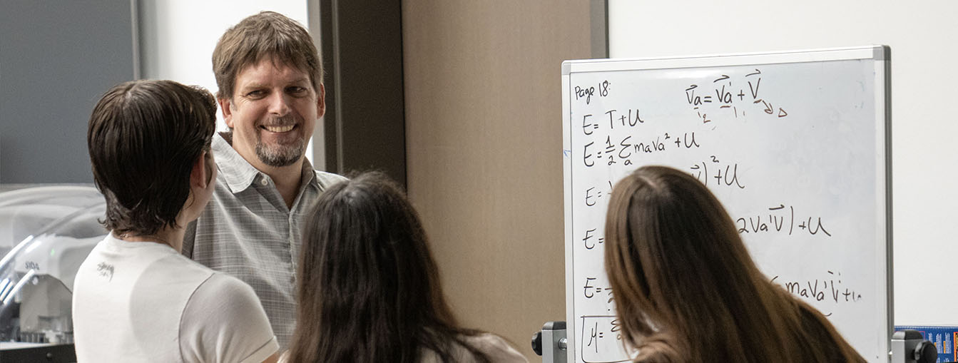 A man smiles at students while reviewing equations on a dry-erase board. 