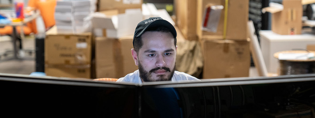 A student in black STARGATE ballcap looks intently at computer screens.