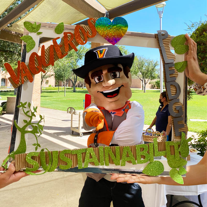 UTRGV Vaquero pointing to the viewer to take the Sustainability Pledge behind the Sustainability Pledge Frame on Campus Sustainability Day