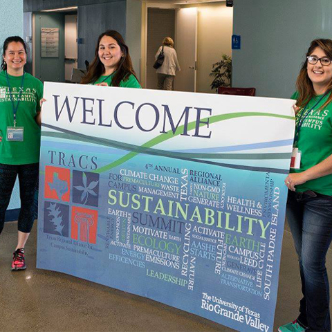 3 females holding up a big welcome sign for the T.R.A.C.K.S convention that UTRGV hosted a couple years ago and on it is the tracks logo, the name of the university, and sustainability words with the word sustainability being in the middle and the biggest in color green