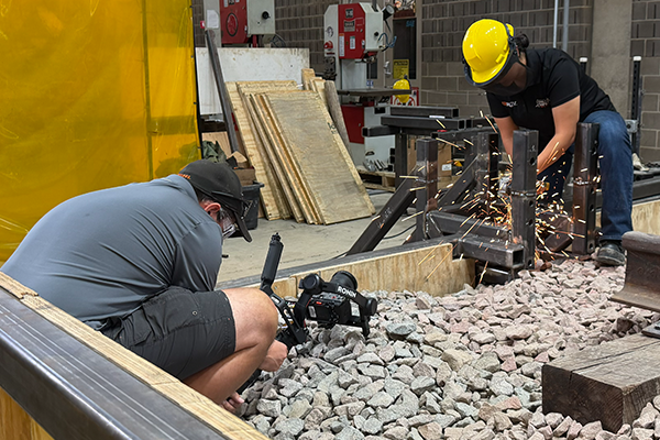 Photo of videographer angling camera upwards to capture an engineering beam being sawed.