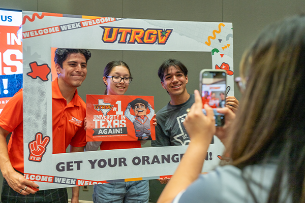 Photo of UTRGV students being photographer with a cellphone while holding a UTRGV social media frame.